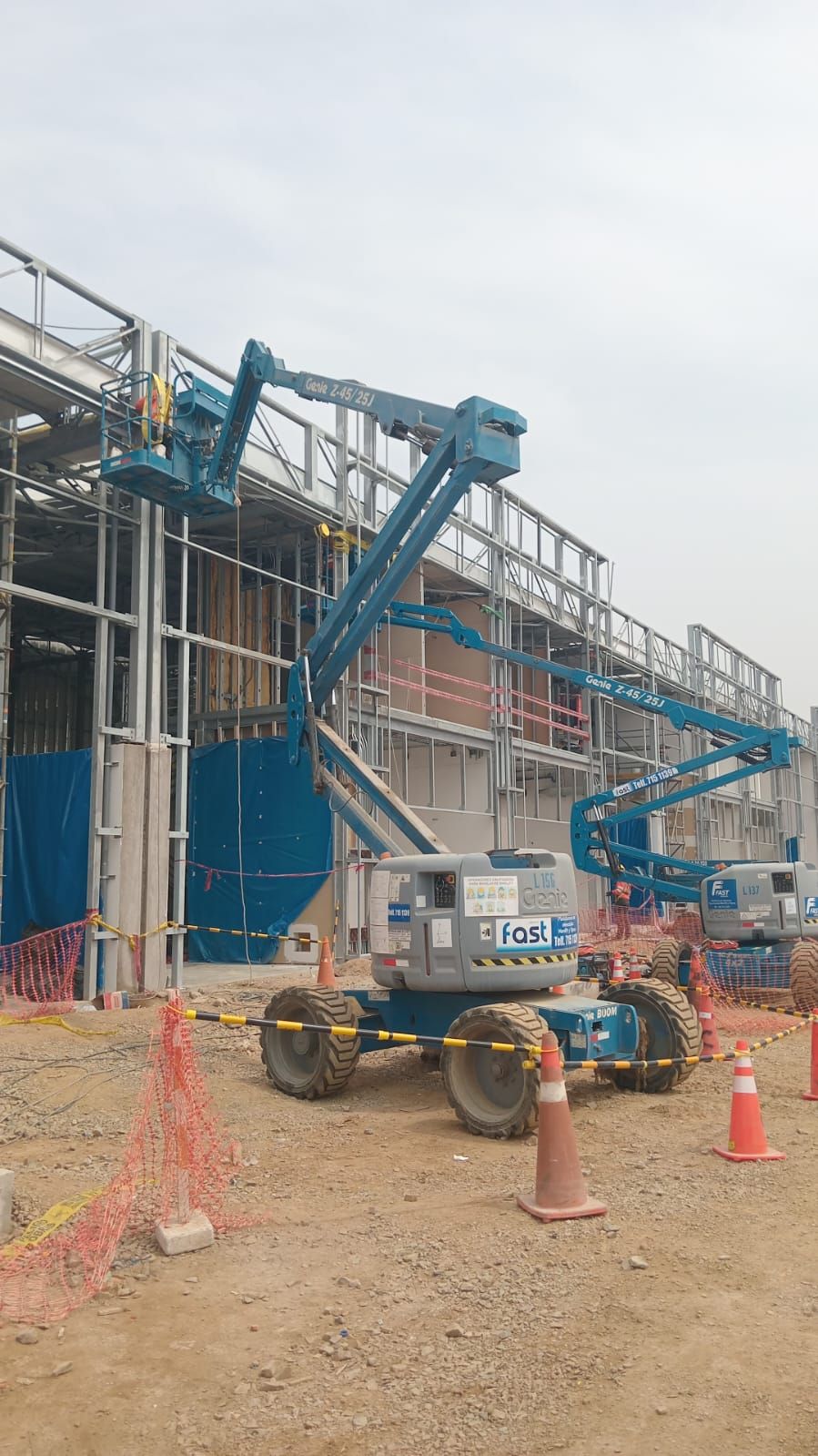 Plataforma elevadora azul junto a andamios en una obra en construcción, con conos naranjas y suelo de tierra.