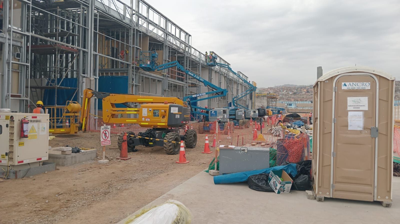 Obra en construcción con trabajadores, maquinaria amarilla, andamios, conos naranjas y un baño portátil.