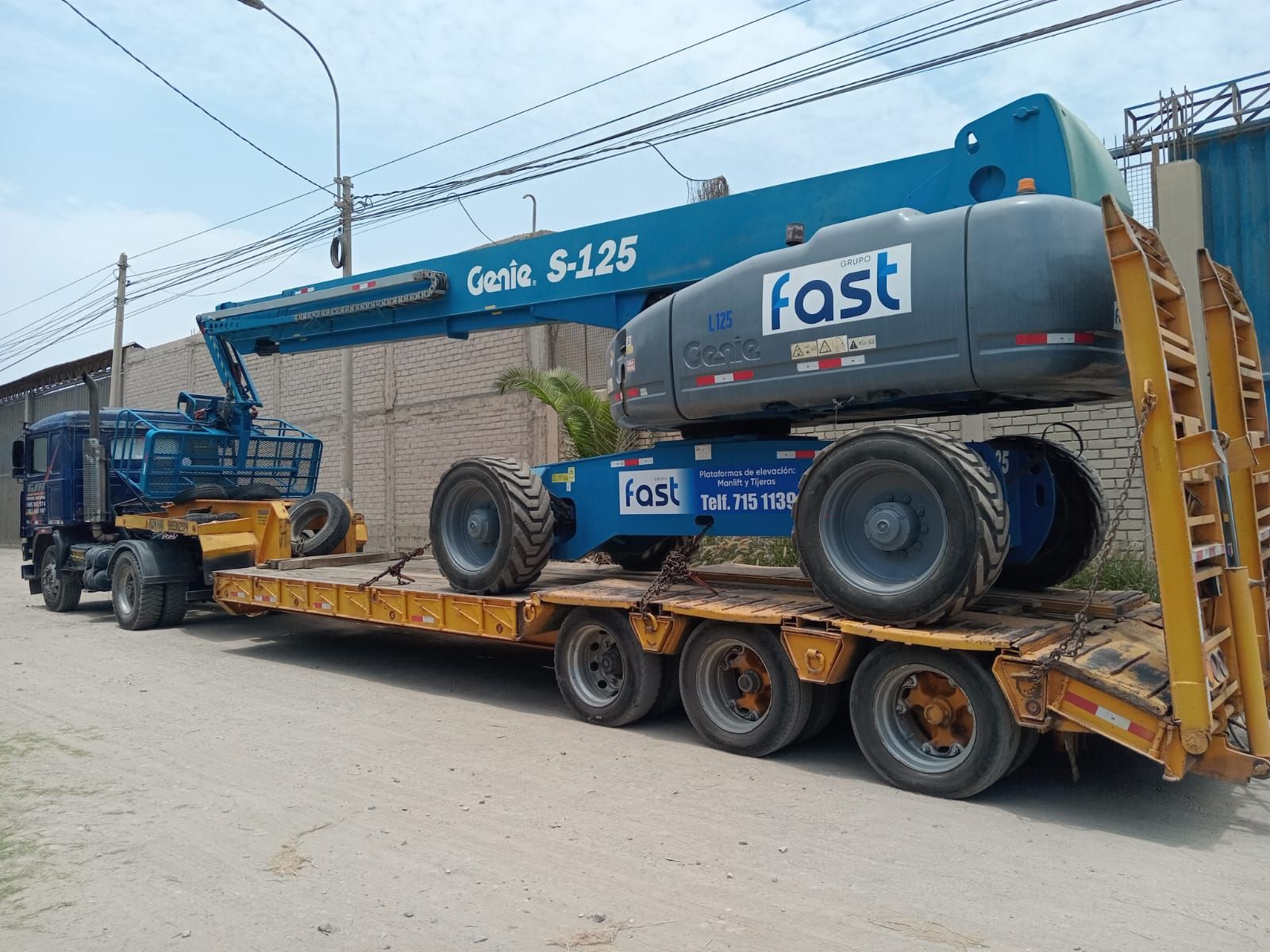 Camión grúa Blue Fast cargado sobre un remolque de plataforma, estacionado en un camino de tierra.