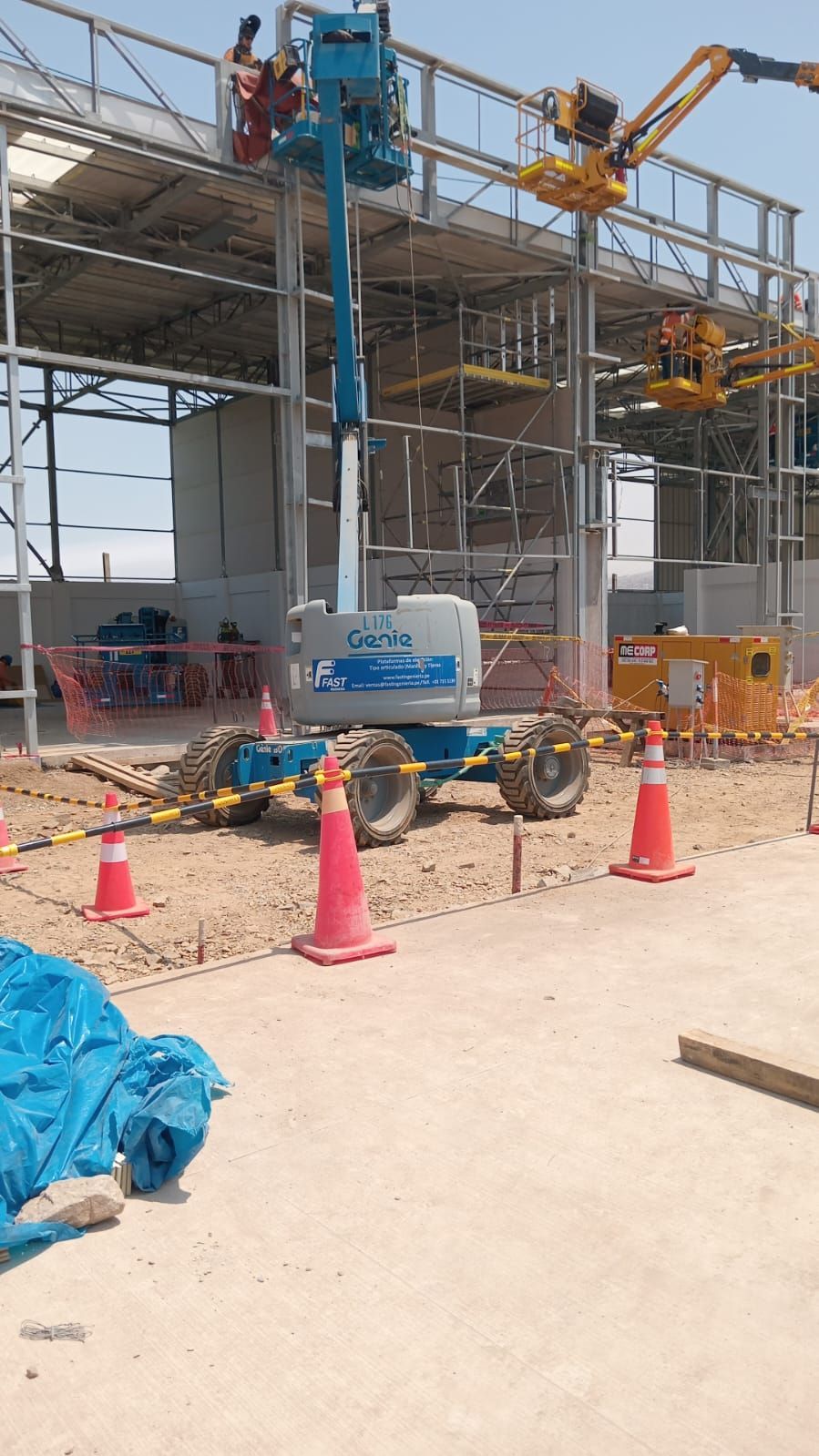 Obra en construcción con andamios, grúas, conos naranjas y un carro de equipo azul junto a una estructura de hormigón.
