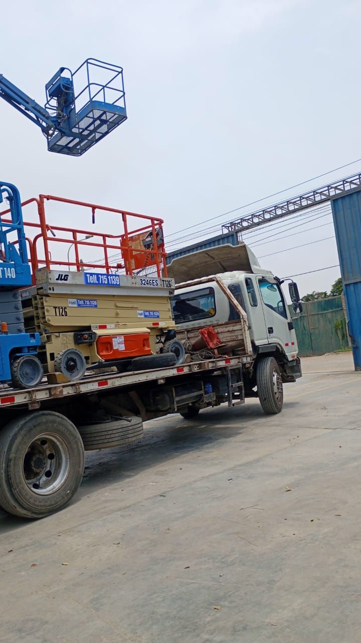 Camión de plataforma cargado de equipo en un patio industrial, con una grúa azul en lo alto.
