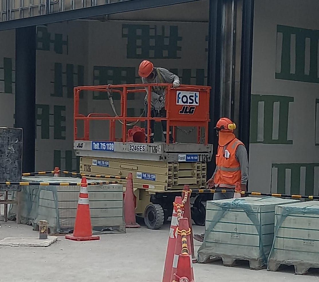 Obrero de la construcción junto a una carretilla elevadora roja y conos naranjas en la entrada de un edificio.