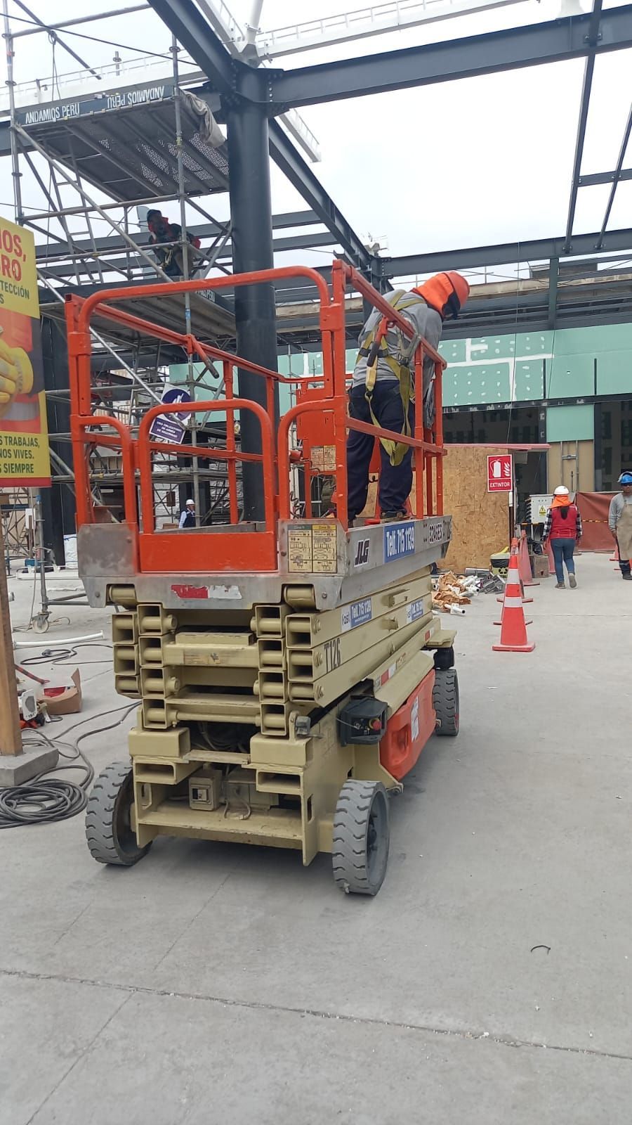Trabajador en una plataforma elevadora de tijera naranja en una zona industrial cubierta con conos de tráfico cerca.