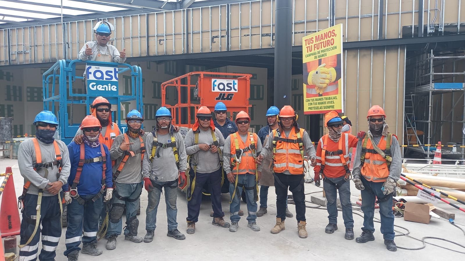 Obreros de la construcción con cascos y chalecos naranjas posando en una obra con maquinaria detrás.