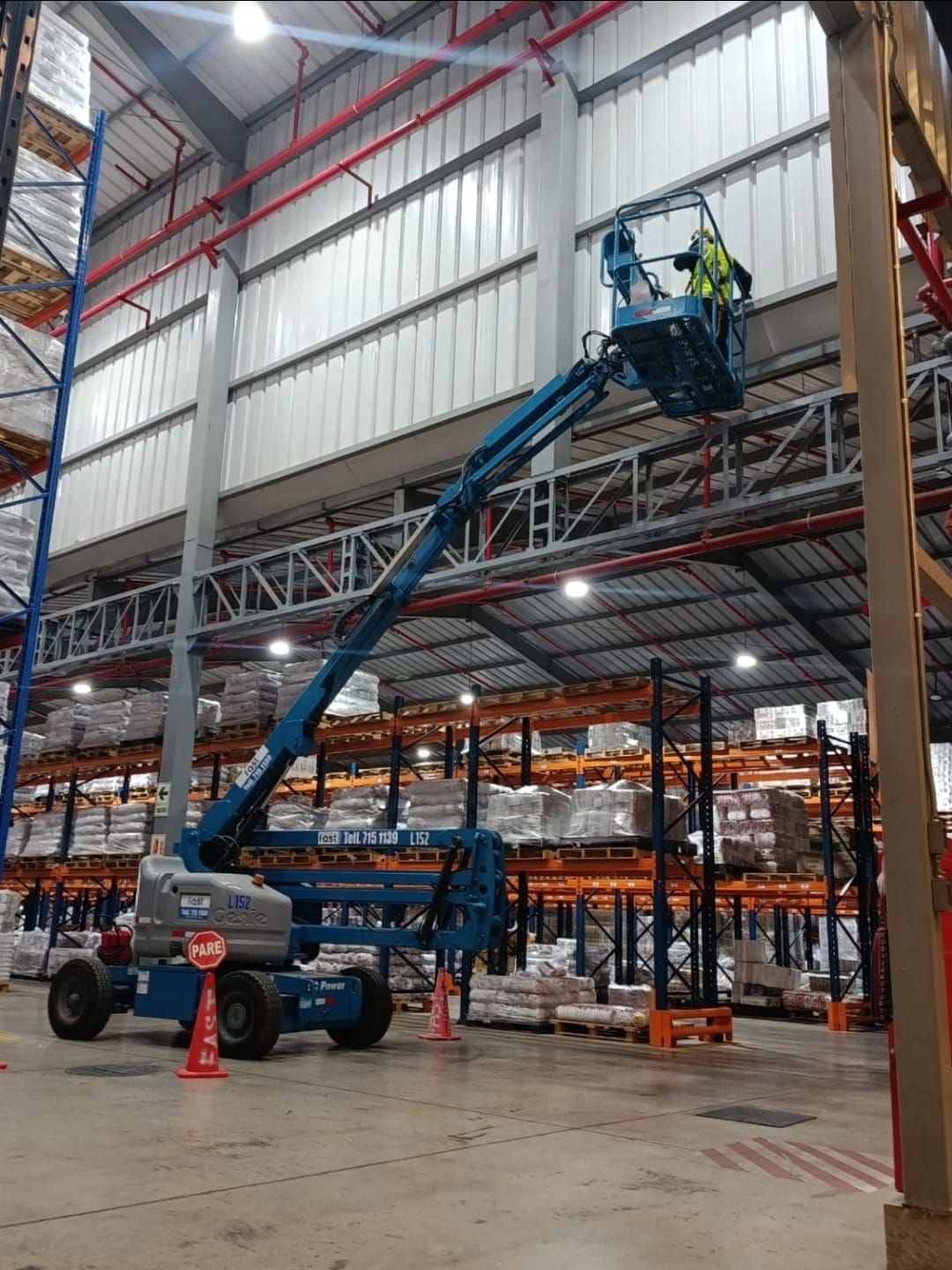 Plataforma elevadora azul con un trabajador en el pasillo del almacén cerca de estanterías naranjas y un techo metálico alto.