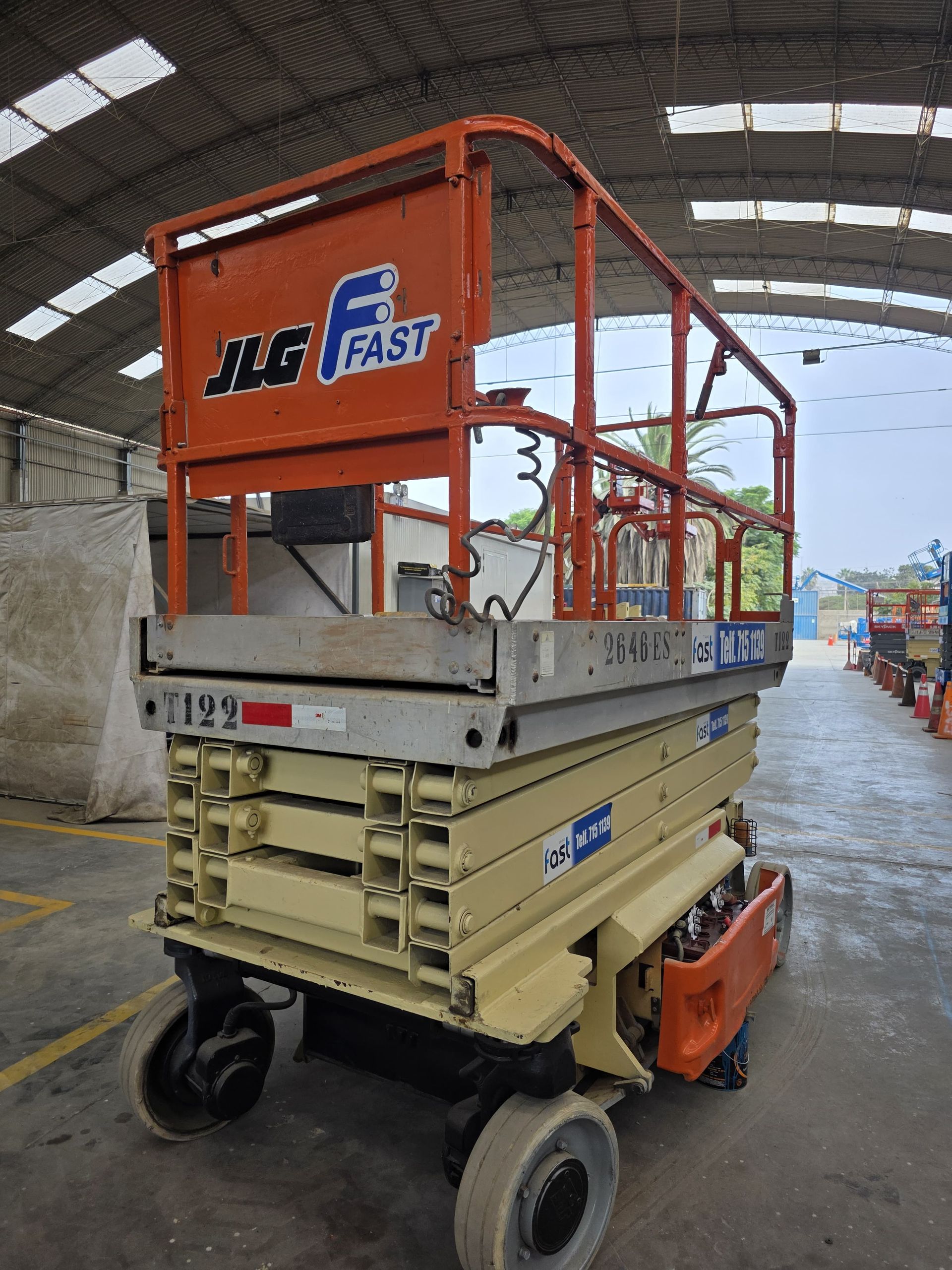 Plataforma elevadora de tijera JLG con barandillas naranjas y plataforma beige, estacionada en un pasillo de un almacén.