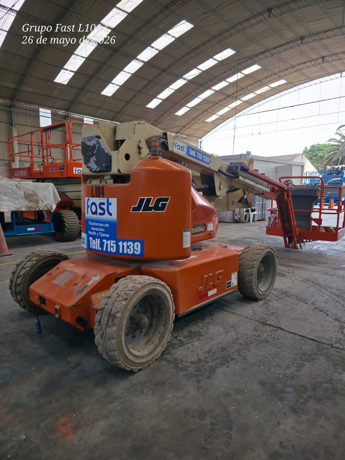 Una plataforma elevadora JLG naranja está estacionada dentro de un almacén con otros equipos de construcción cerca.