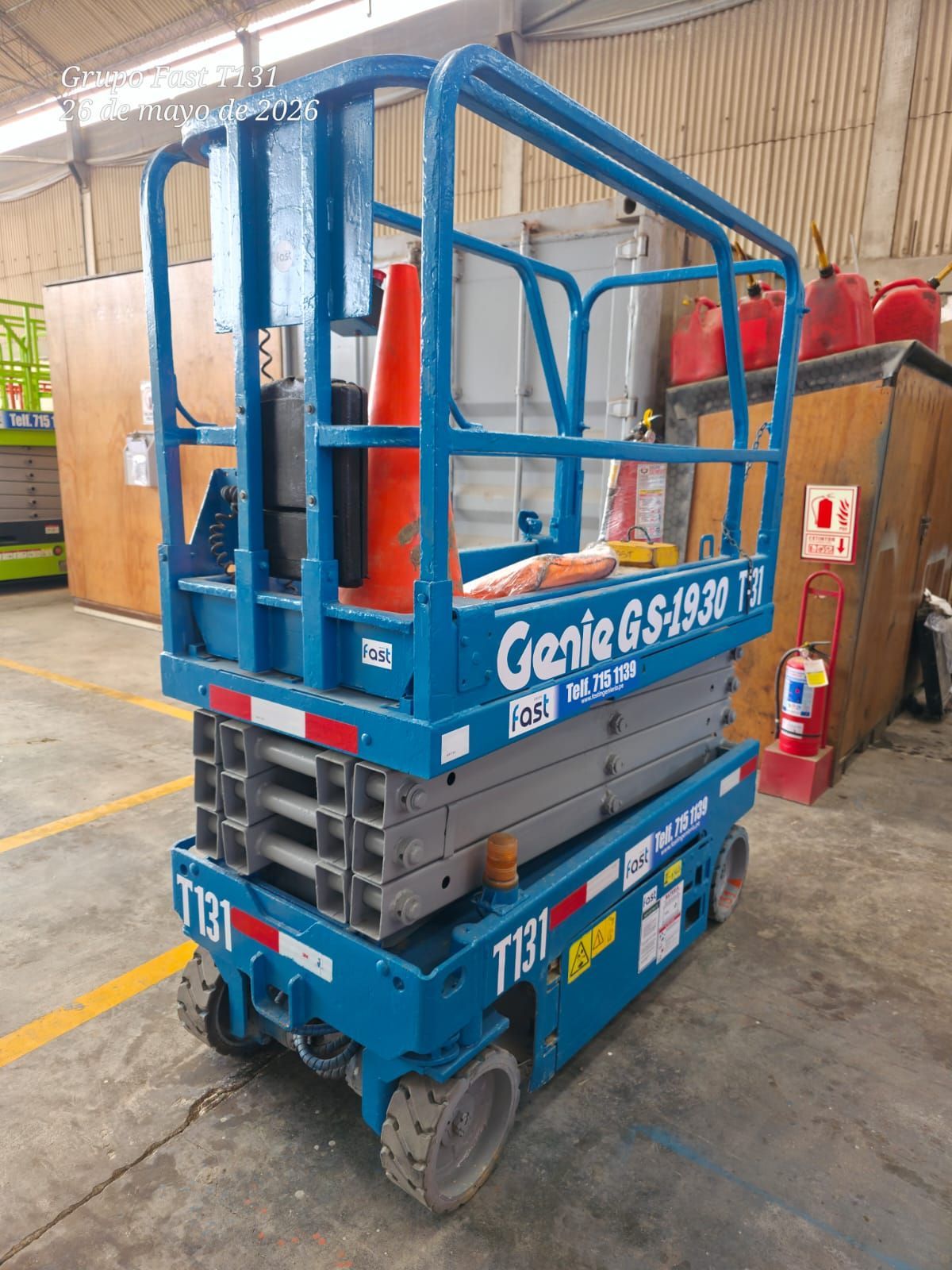 Plataforma elevadora de tijera Blue Genie GS-1930 estacionada en el interior de un almacén, con barandillas de seguridad y conos naranjas en su plataforma.