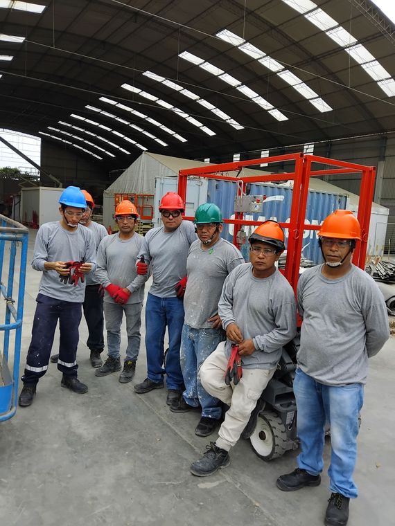 Dos trabajadores con chalecos de seguridad y cascos están de pie junto a una plataforma elevadora de tijera amarilla en un almacén.