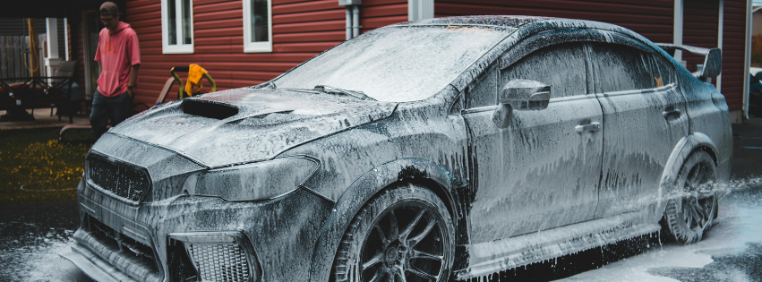 Black sports car covered in thick foam during a car wash, parked outdoors near a red building