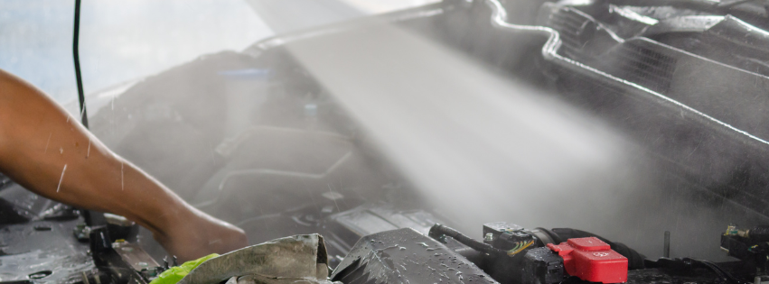 A person cleaning a car engine with a high-pressure water spray, showing steam and water mist 