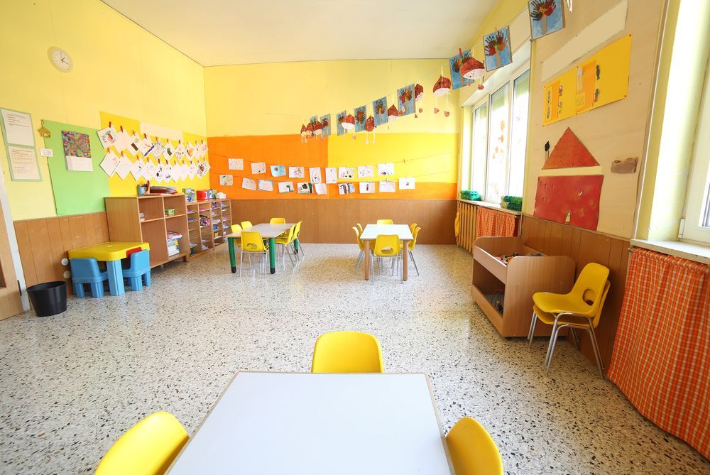 Colorful classroom with small tables, chairs, and children’s artwork on yellow walls.
