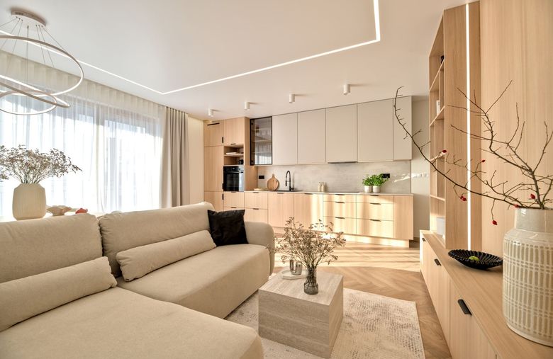 Bright modern living room with beige sectional, wood cabinetry, and open kitchen in soft neutral tones