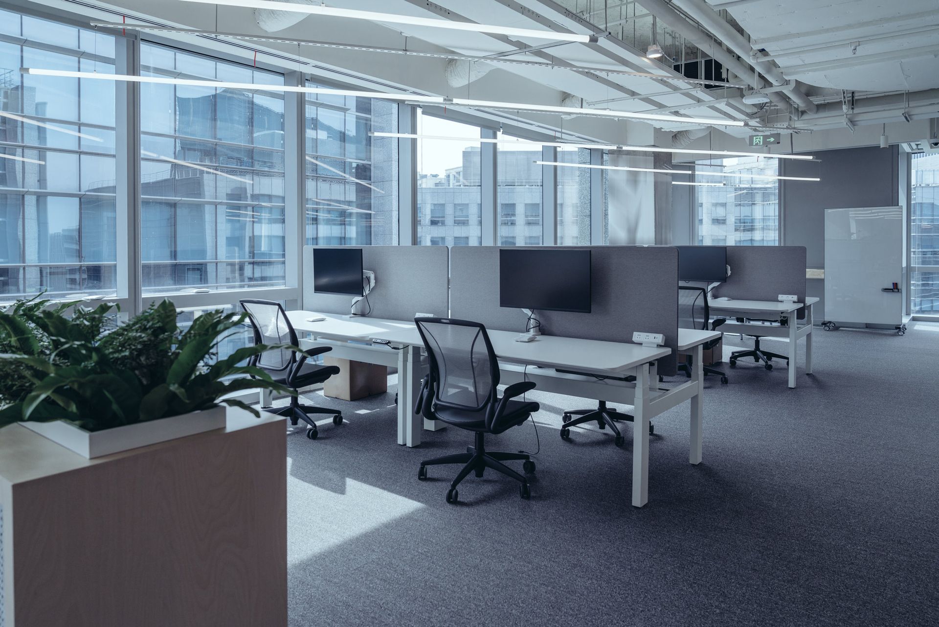 Modern open-plan office with desks, monitors, chairs, and large windows with natural light