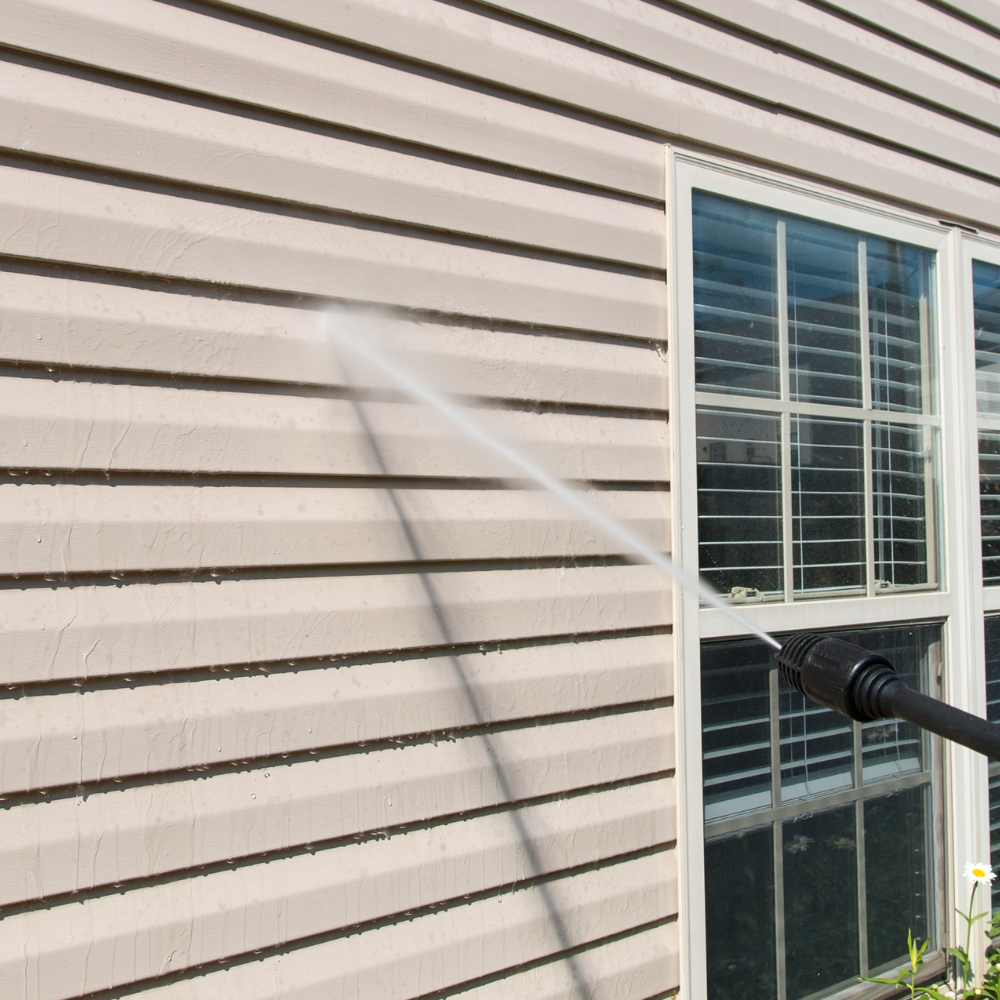 A person power washes siding of a house near a window.