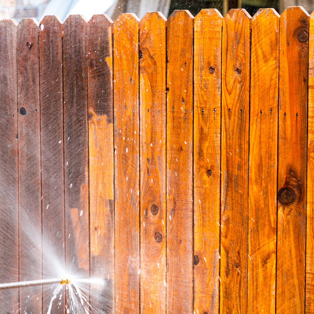 Power washing a stained wooden fence, showing the cleaned vs. dirty sections.