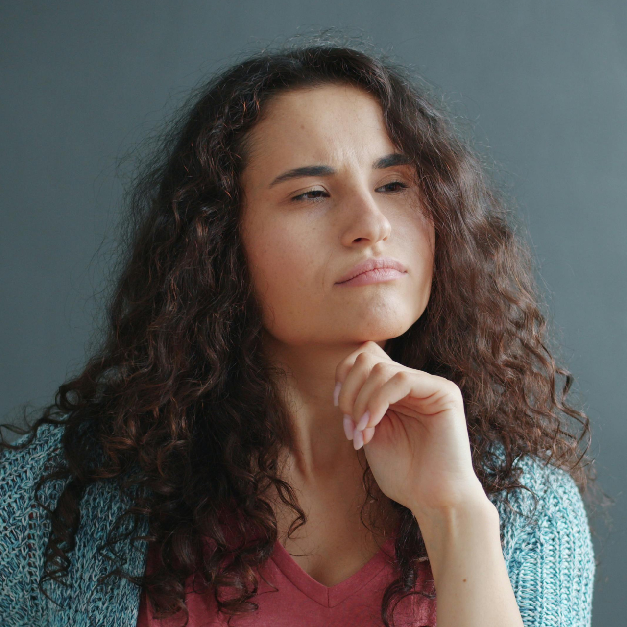 A person with long, curly dark hair looks up thoughtfully with a hand resting against their chin.