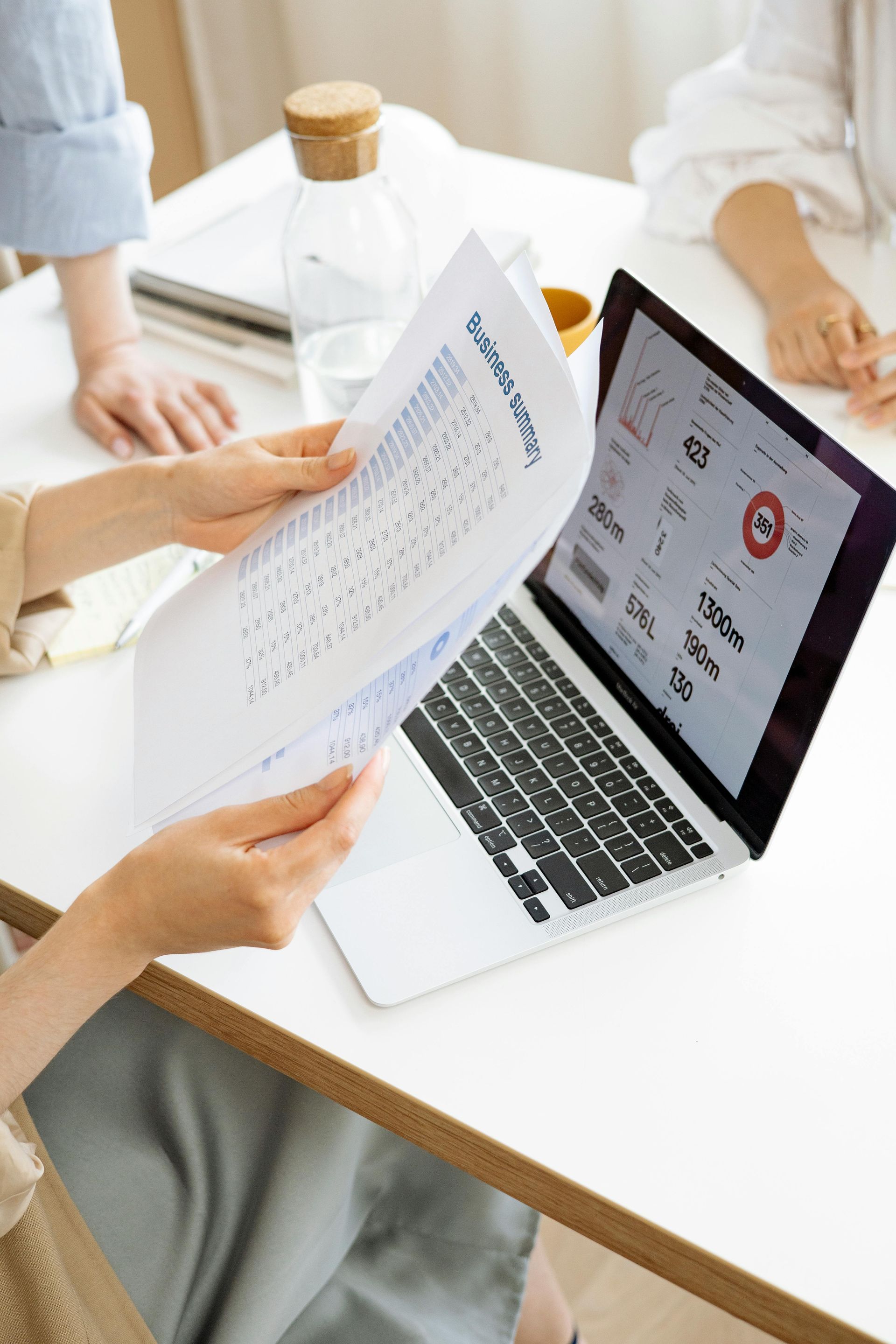 Hands flipping through papers while reviewing laptop data at a table.