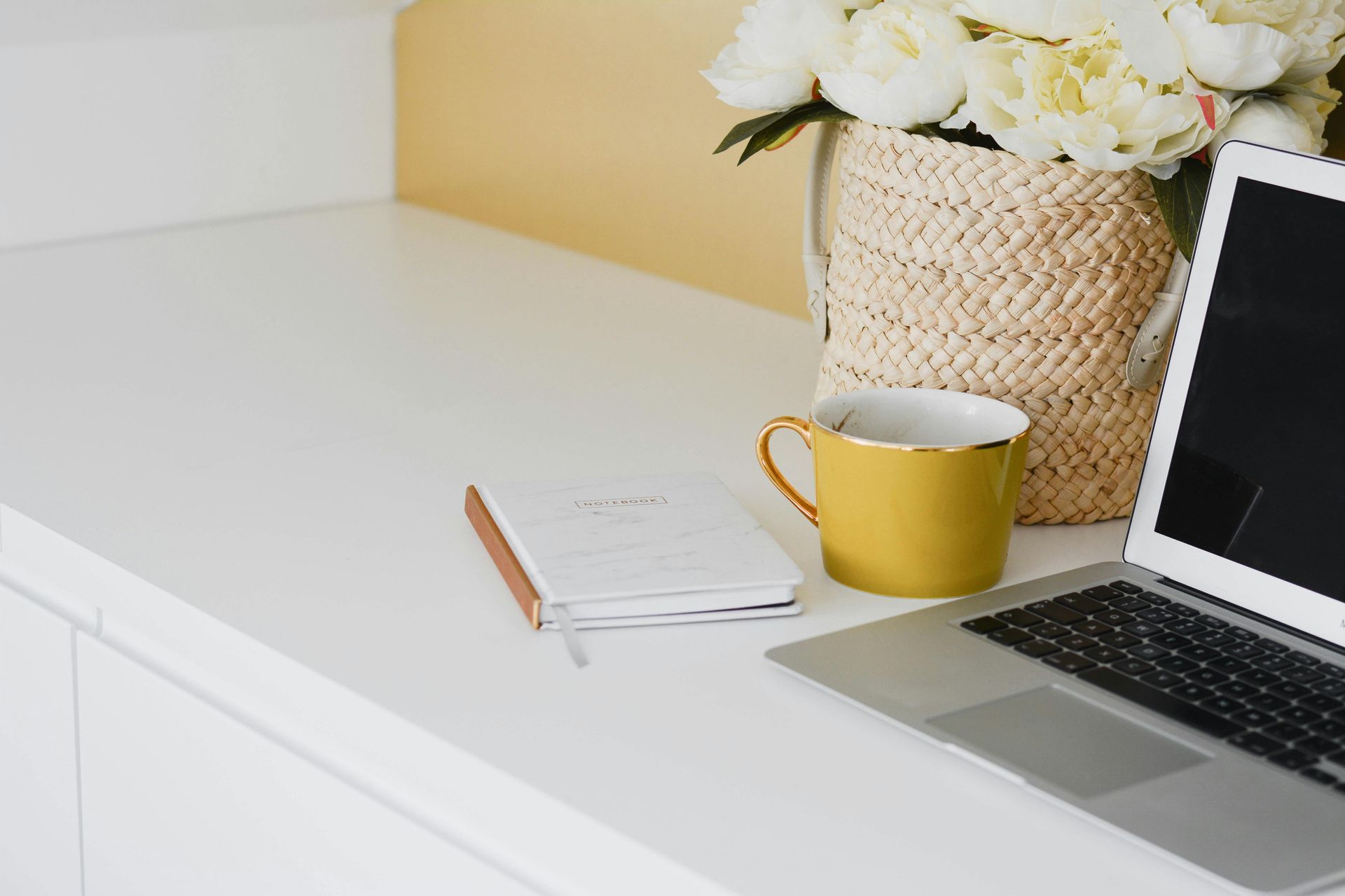 White desk with laptop, yellow mug, flowers in basket, and a notebook.