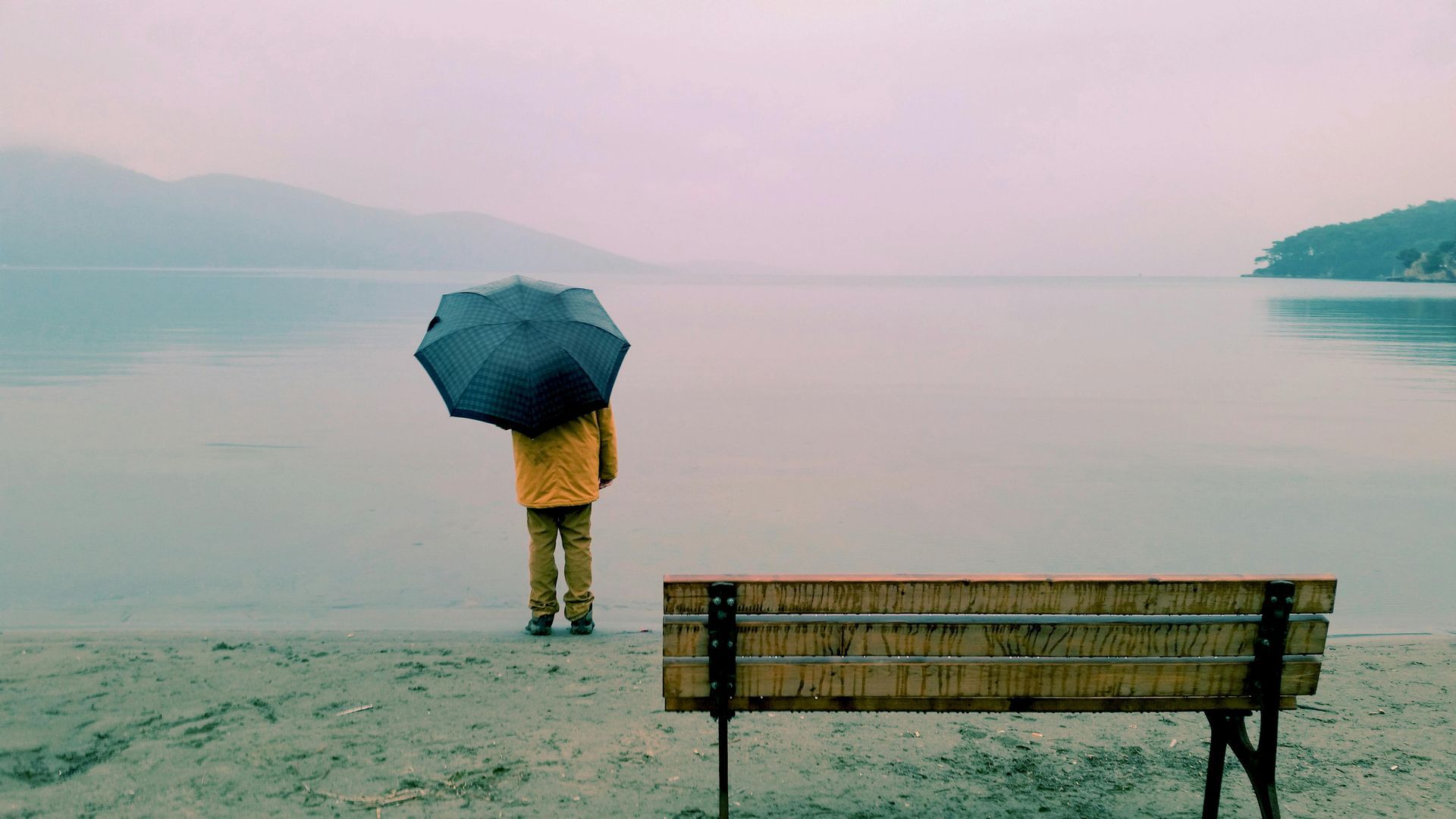 A person holding an umbrella is standing on a beach next to a bench.