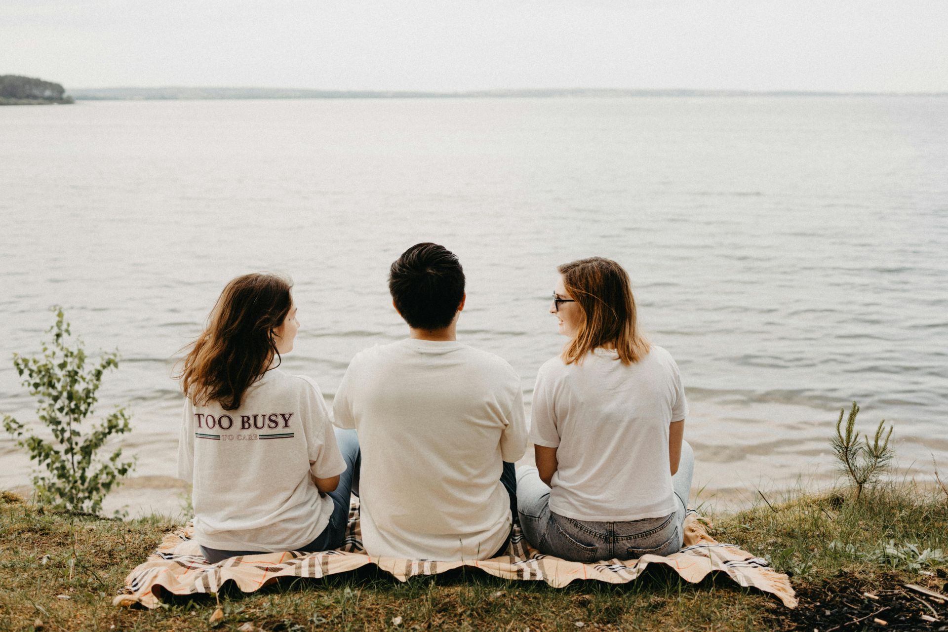 Three people sitting on a blanket by a calm body of water. They are facing the water.