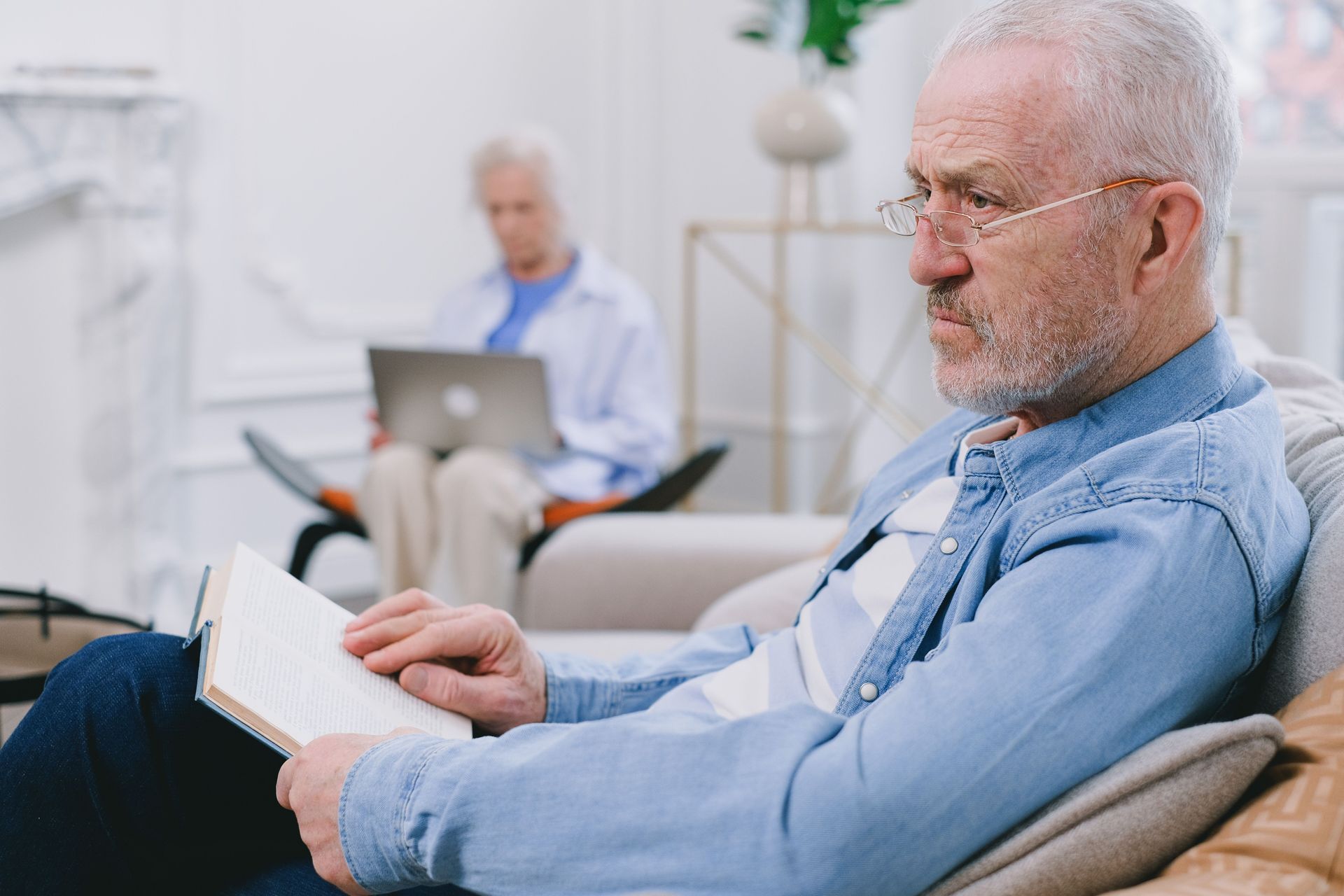 Man reading a book on a couch in a bright living room while another person uses a laptop in the background