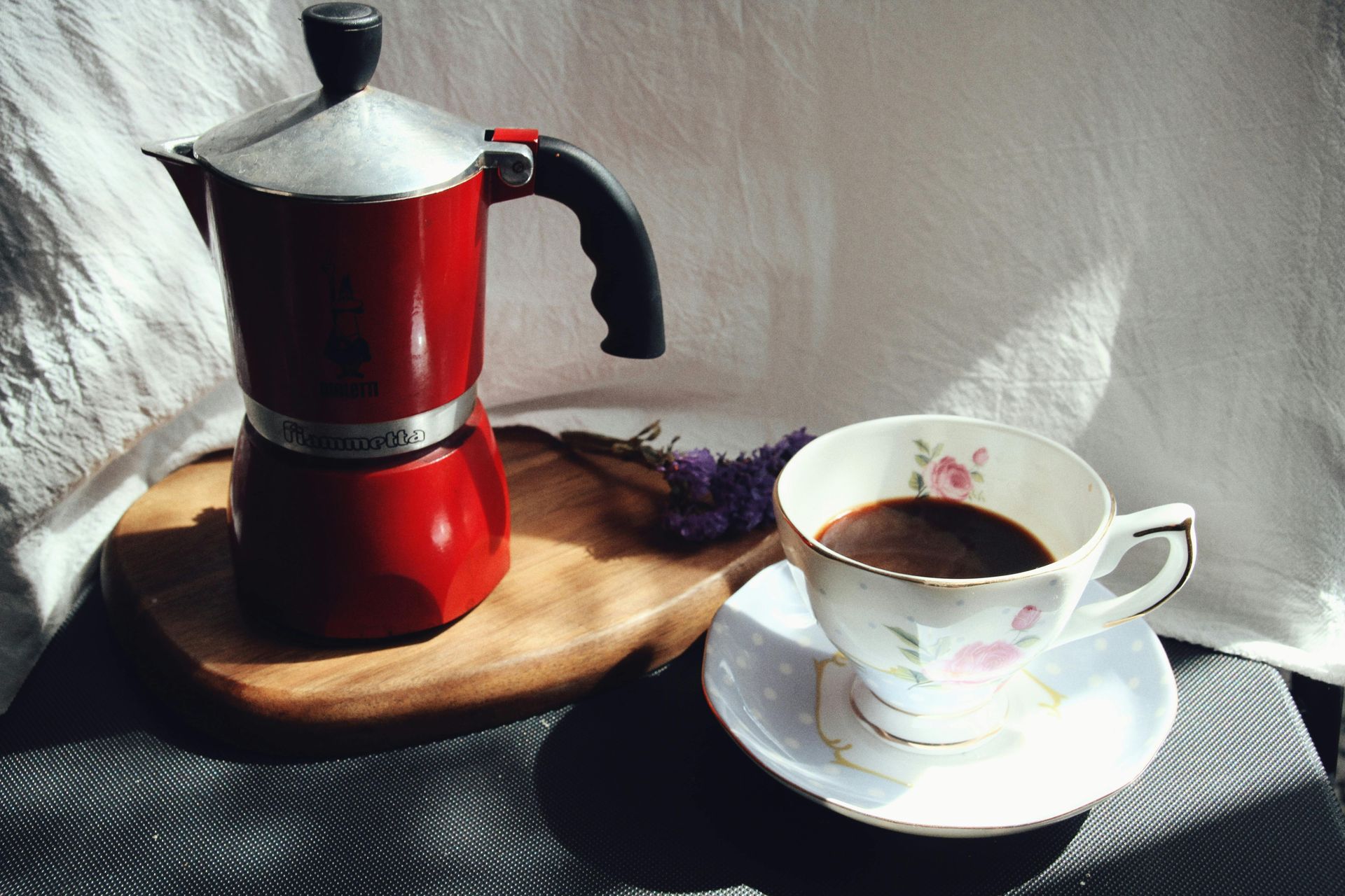 A red stovetop espresso maker sits on a wooden board next to a filled floral teacup and saucer in sunlight.