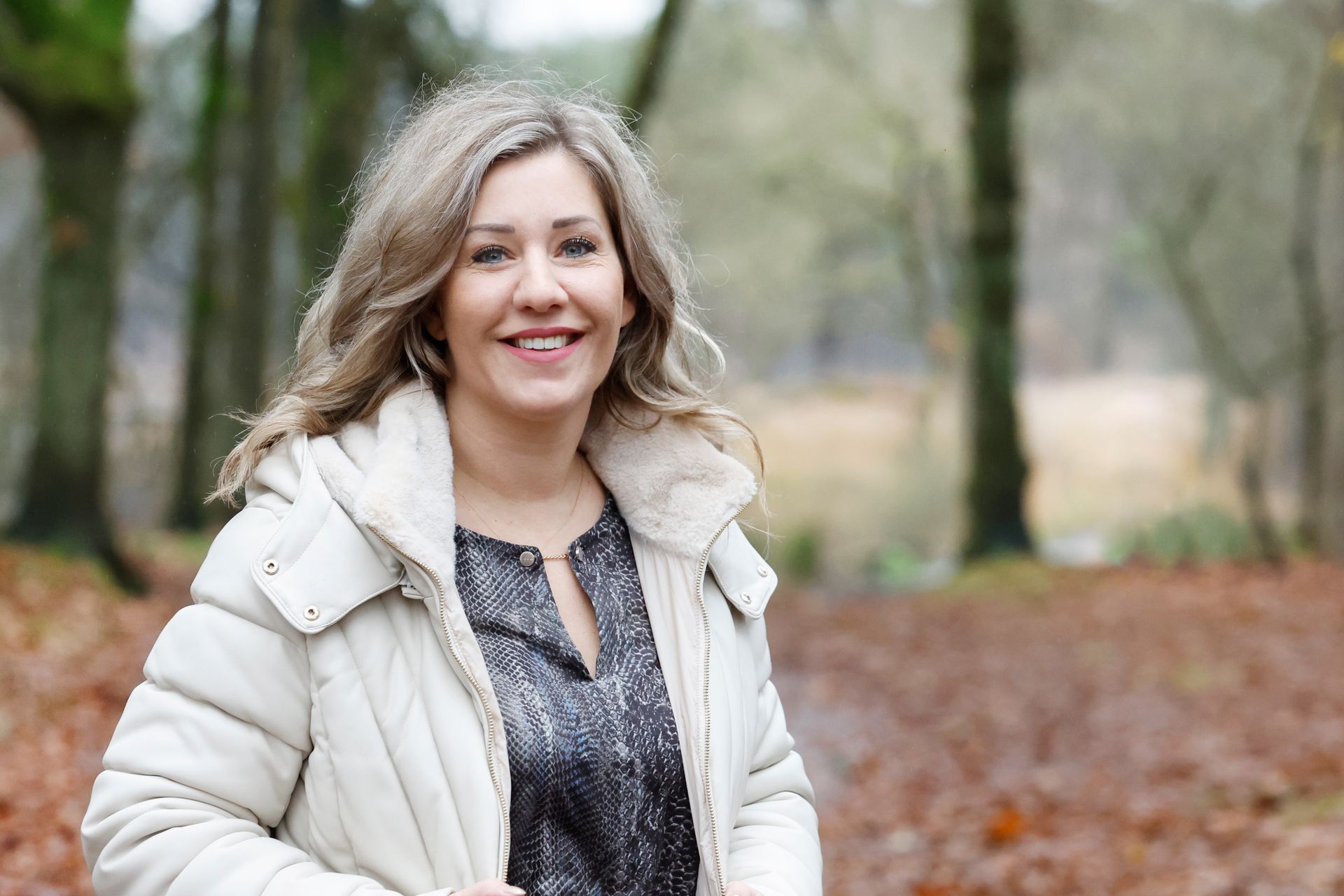 Een vrouw in een wit jasje staat in het bos en lacht.