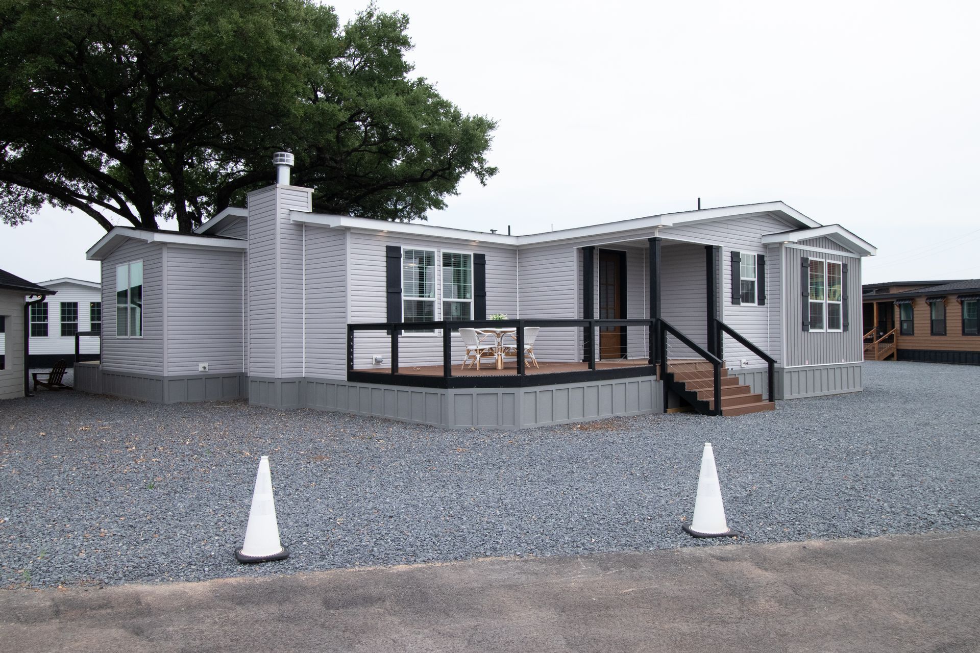 Small gray modular home with a gravel yard and cone markers under a cloudy sky
