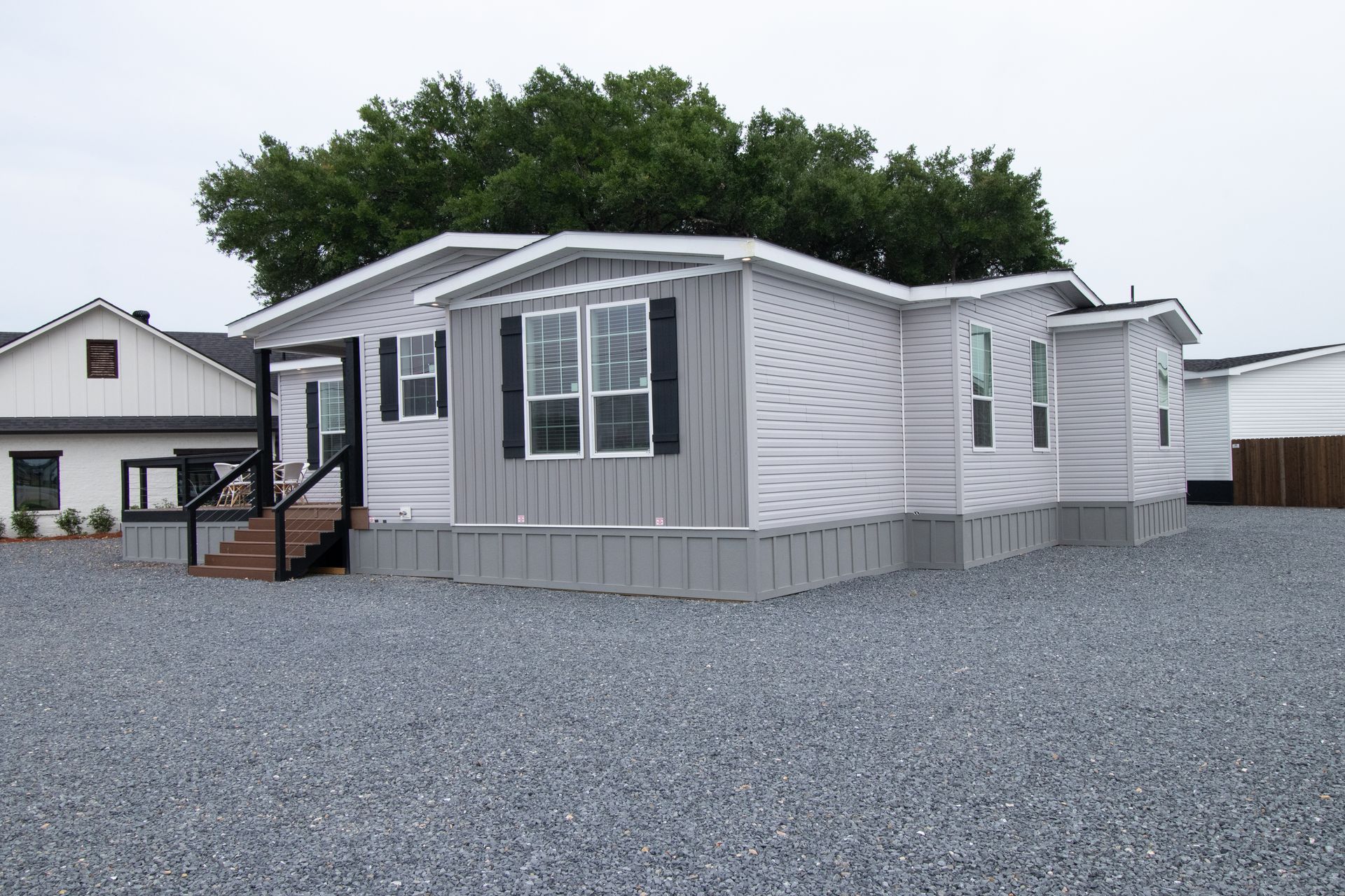 Gray manufactured home with front steps and gravel lot, parked beside another house and trees