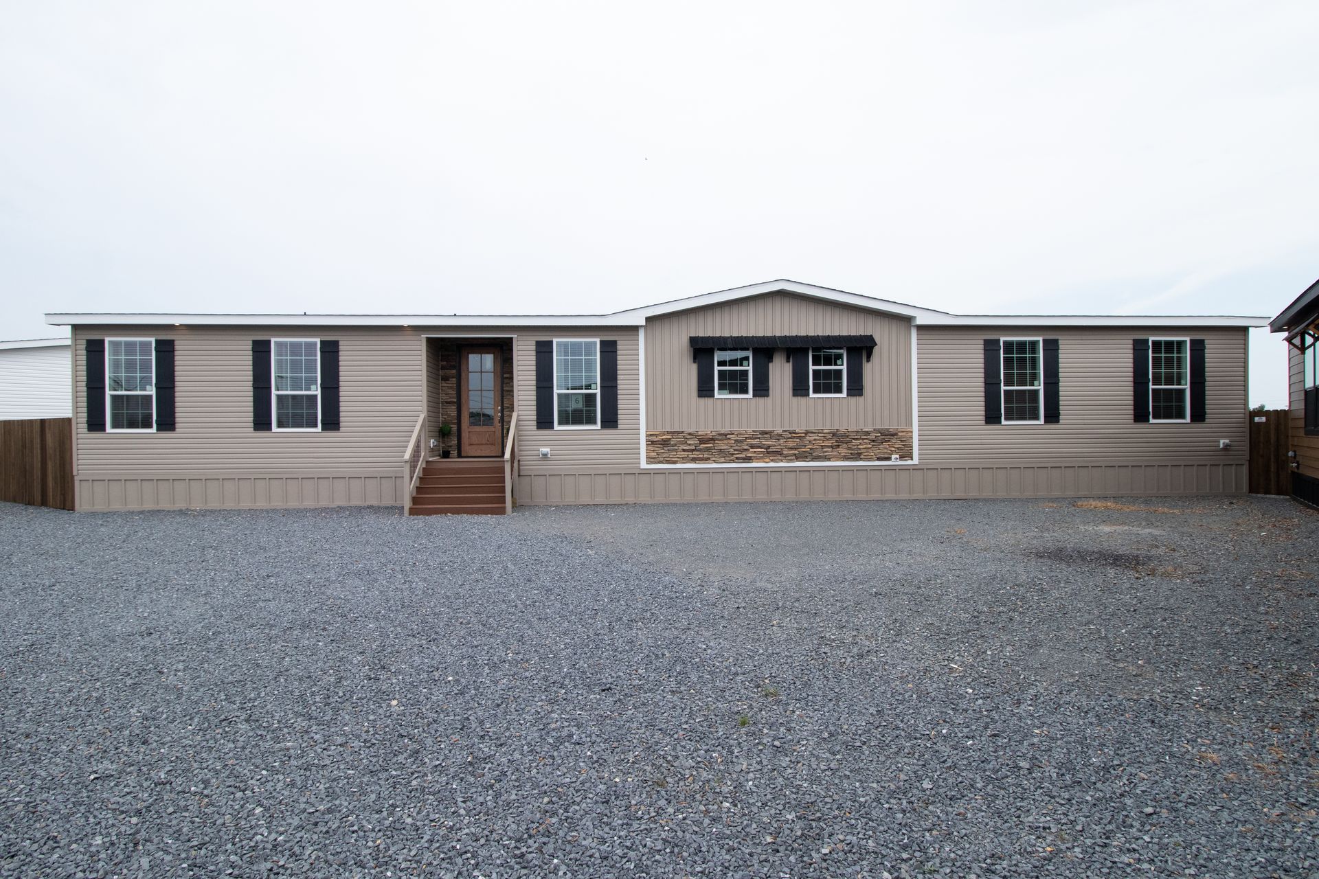 Single-story beige mobile home with a central porch, set on a gravel lot under a cloudy sky.