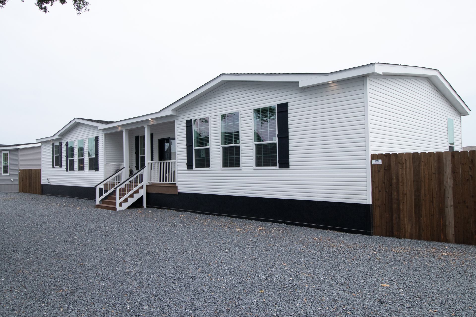 White manufactured home with porch and stairs on a gravel lot under an overcast sky