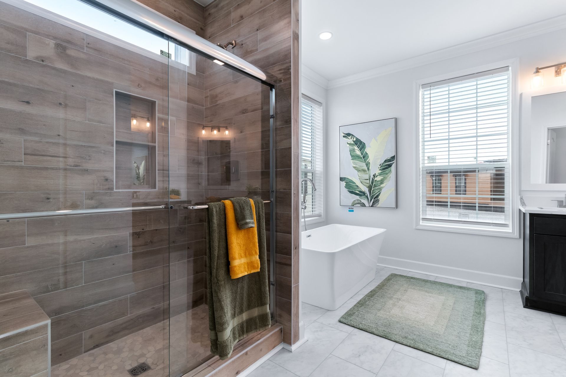 Modern bathroom with glass shower, white tub, gray rug, and yellow towels