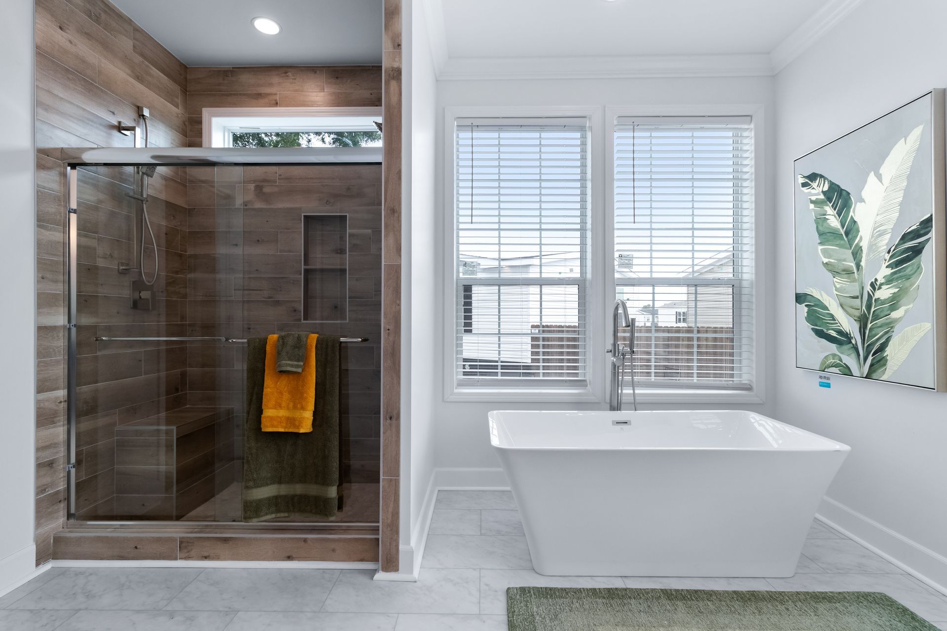 Modern bathroom with glass shower, freestanding tub, and large windows with white blinds
