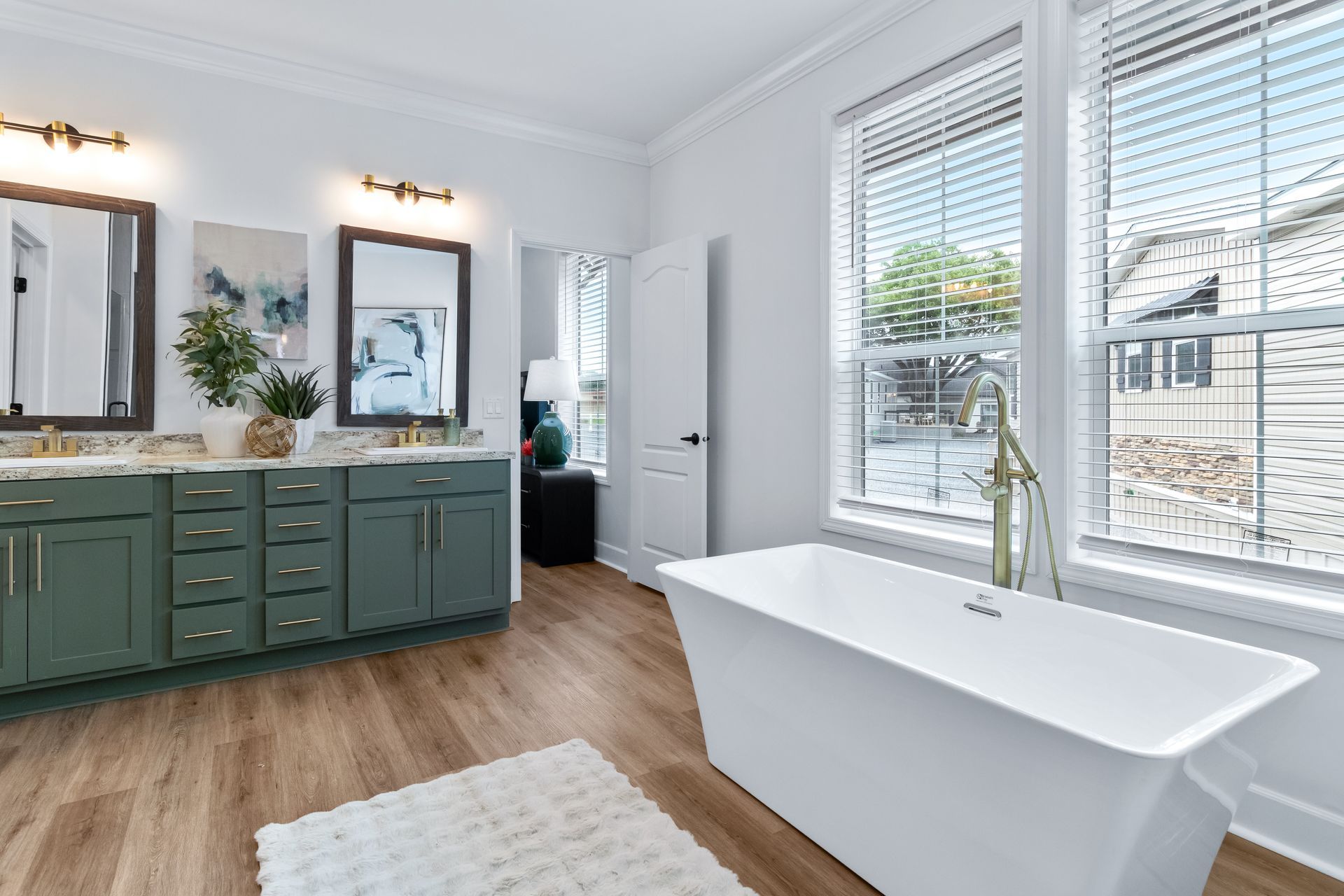Bright bathroom with green vanity, double mirrors, wood floor, and a freestanding tub by large windows