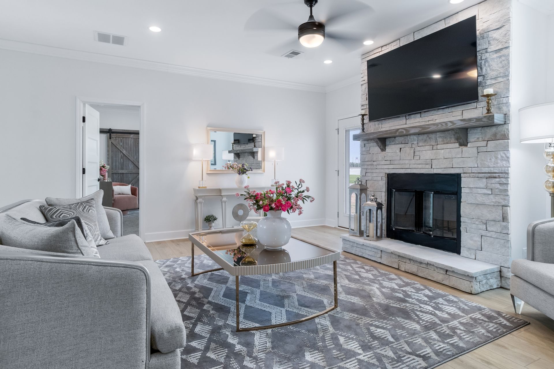 Bright living room with gray sofa, patterned rug, stone fireplace, wall-mounted TV, and gold coffee table.