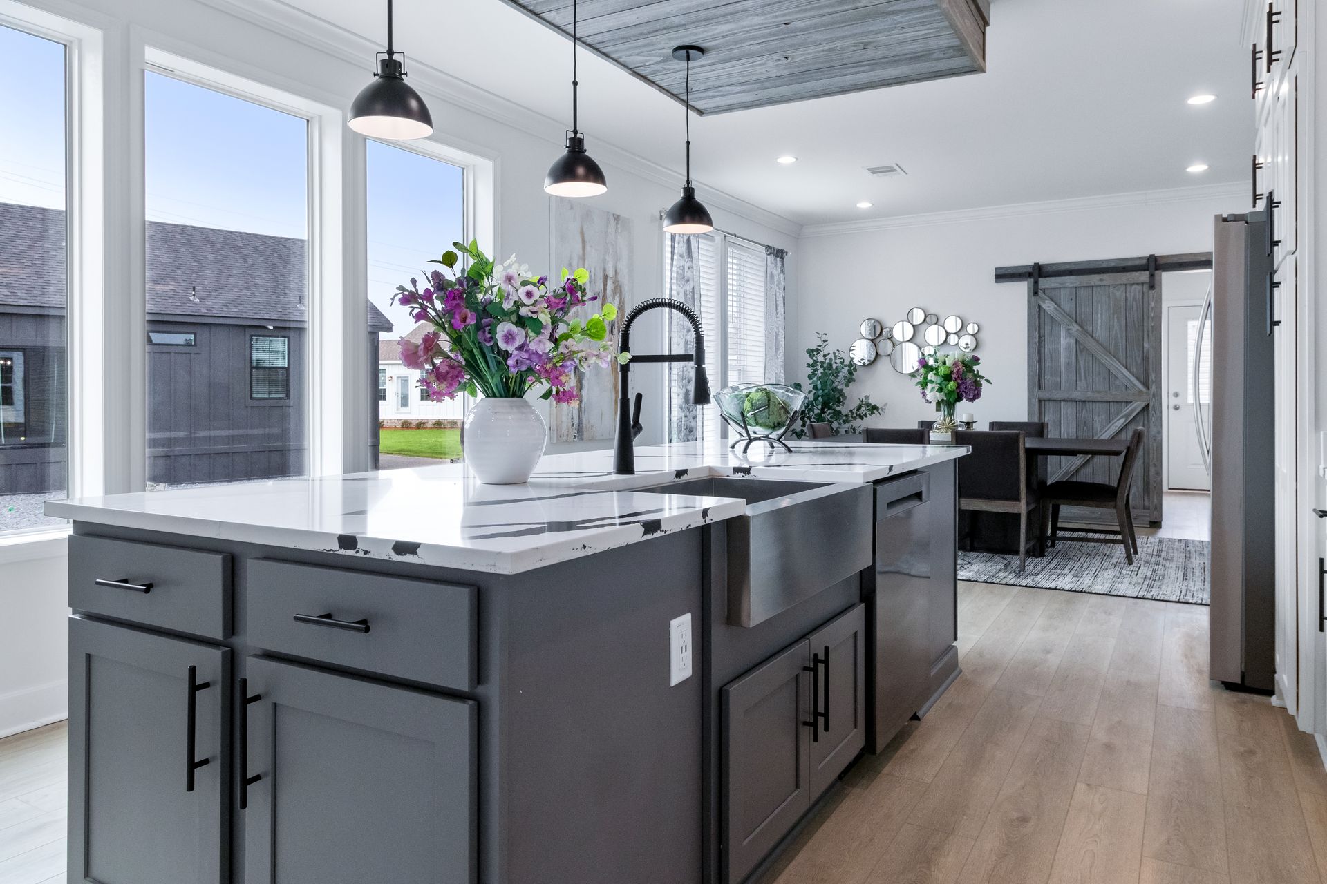 Modern kitchen island with gray cabinets, white countertops, pendant lights, and flowers, opening to a dining area