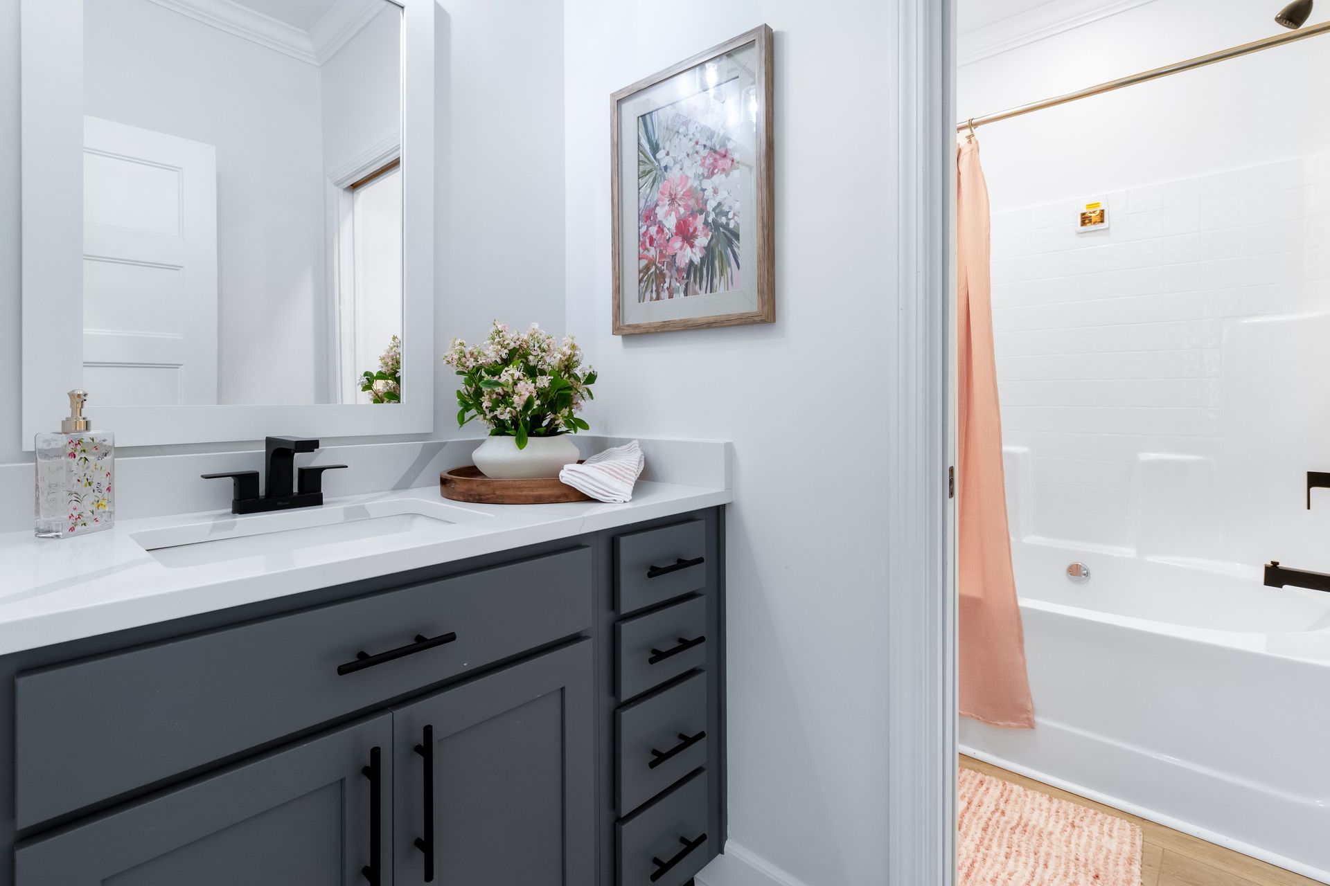 Bright bathroom vanity with mirror, flowers, and an open view into a tiled shower with peach curtain
