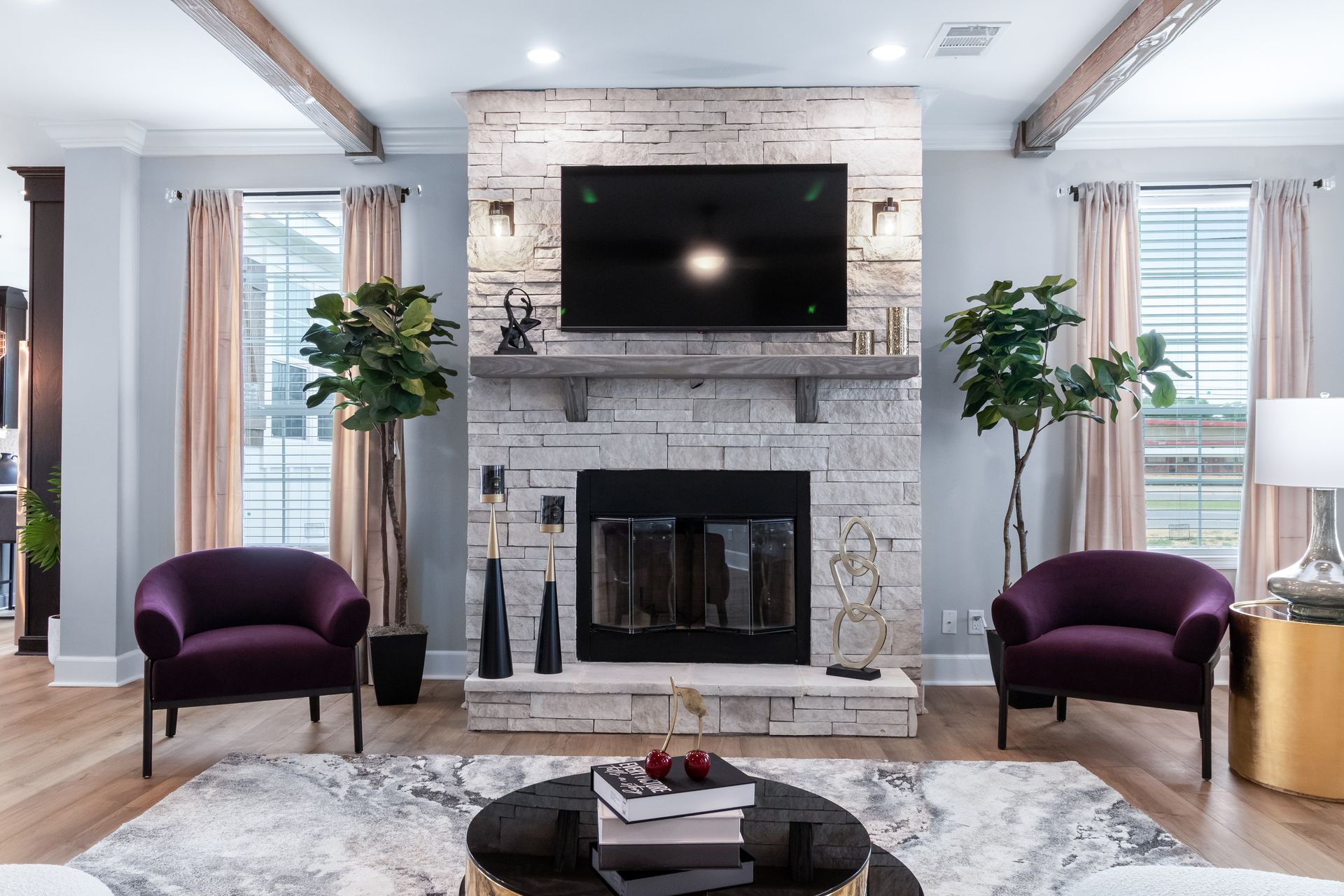 Bright living room with stone fireplace, wall-mounted TV, and two purple chairs by tall windows.