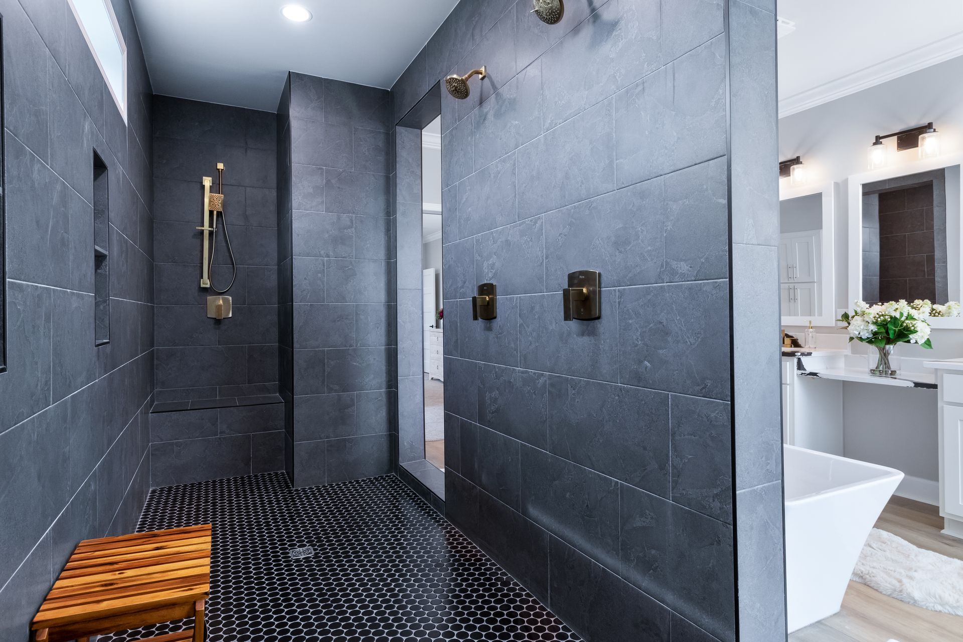 Modern gray tiled shower with black floor, brass fixtures, and a wooden stool in a sleek bathroom