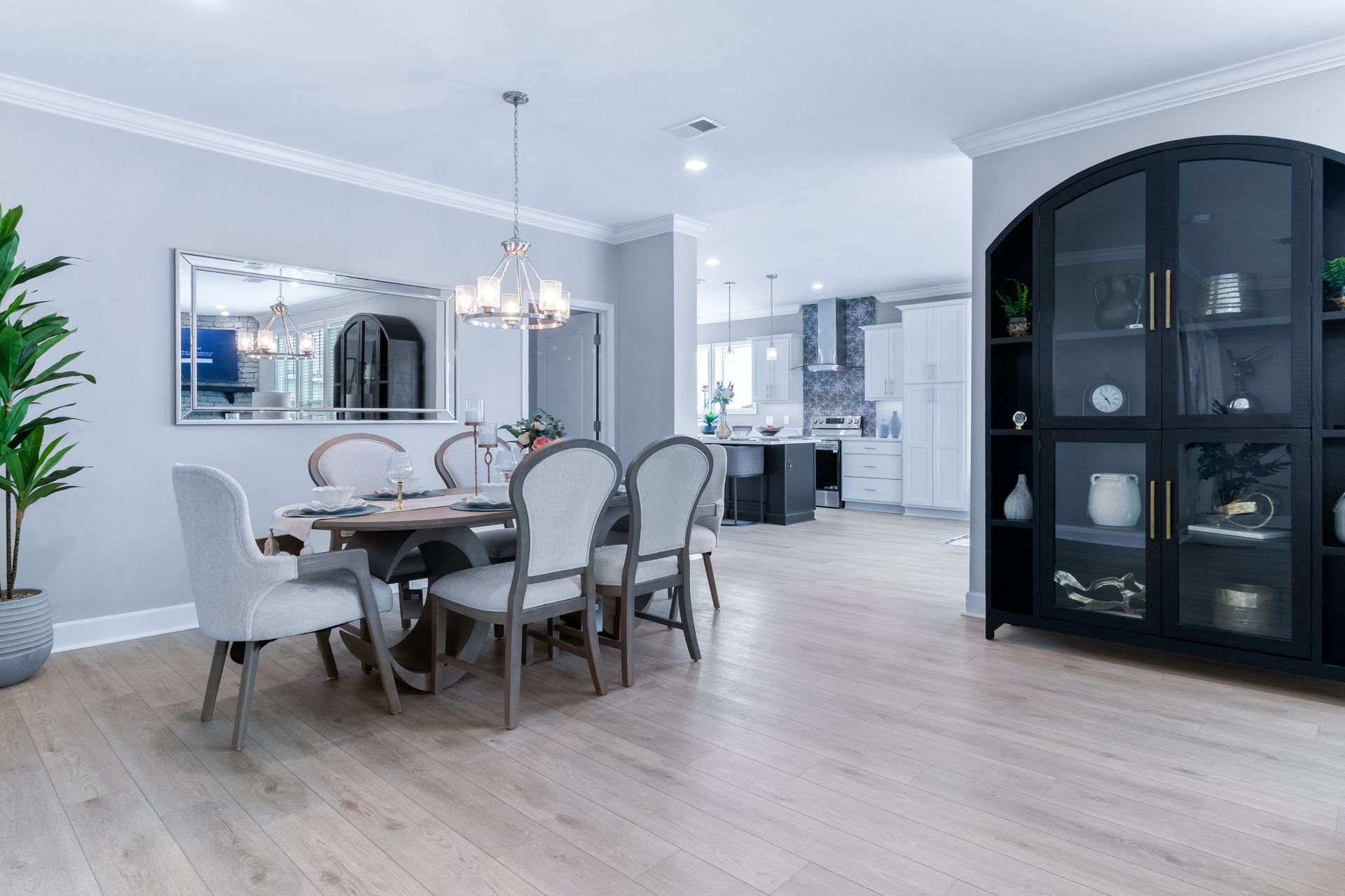 Bright modern dining room with a glass table, white chairs, chandelier, and dark arched cabinet by large windows