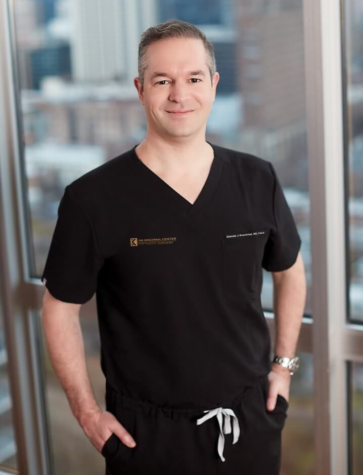 Woman with long dark hair, smiling, in front of a wall of colorful flowers, wearing a black blazer.