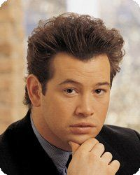 Man with brown hair, in a black jacket, resting chin on hand. Soft lighting in a blurred setting.