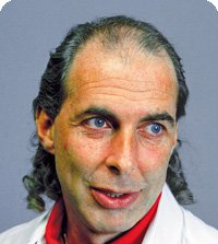 Man with receding hairline, blue eyes, and curly sideburns smiling; wearing a red shirt and white coat.