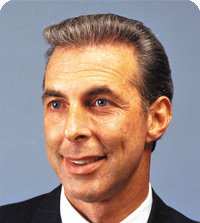 Man with short, graying hair and blue eyes, smiling, wearing a dark suit.