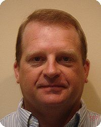 Man with short brown hair, looking directly at the camera. Wearing a light blue and white striped shirt.