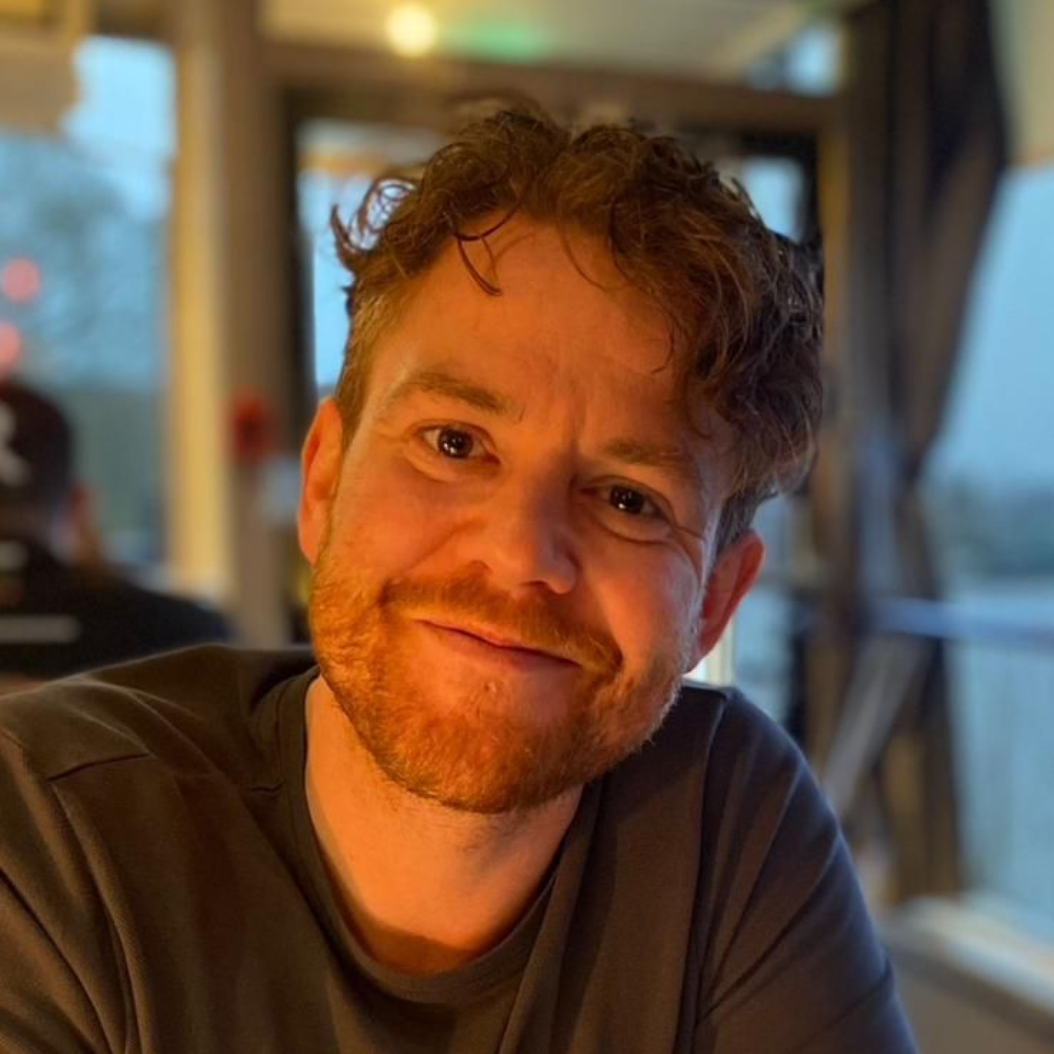 Man with curly hair wearing a black t-shirt smiles in front of green foliage.