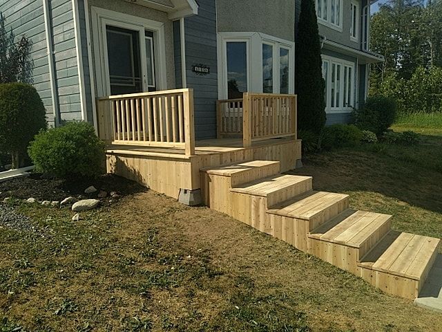 Newly built wooden deck and stairs attached to the side entrance of a grey-sided house, set in a yard with green grass.