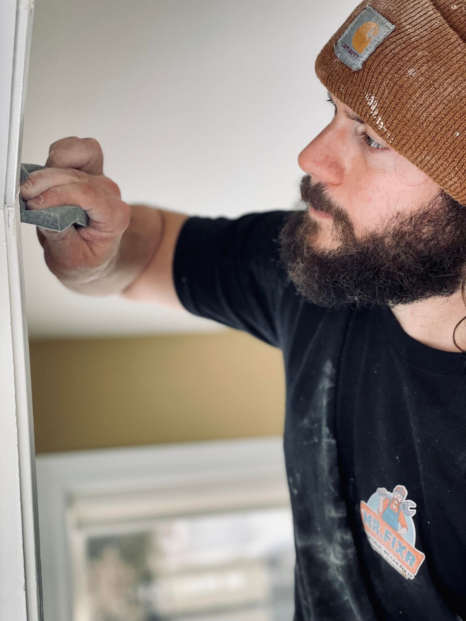 A person wearing a brown knit beanie and black shirt uses a small sanding block to smooth a wall edge.