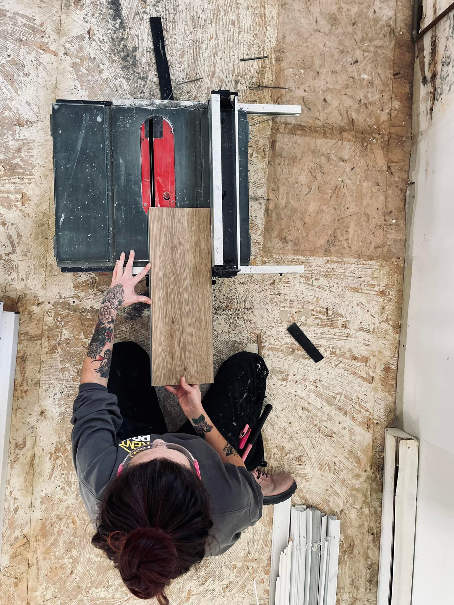 An overhead view of a person using a table saw to cut a plank of wood flooring in a workshop.